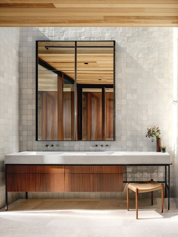 A refined contemporary bathroom featuring a long floating timber vanity with integrated double basins, a pale stone benchtop, and a large black-framed mirrored cabinet. The walls are clad in small textured light-grey tiles, while warm timber ceiling boards and rich timber cabinetry create contrast. A woven-seat stool and small vase add understated softness. The mirror reflects timber-lined surfaces and black structural detailing.