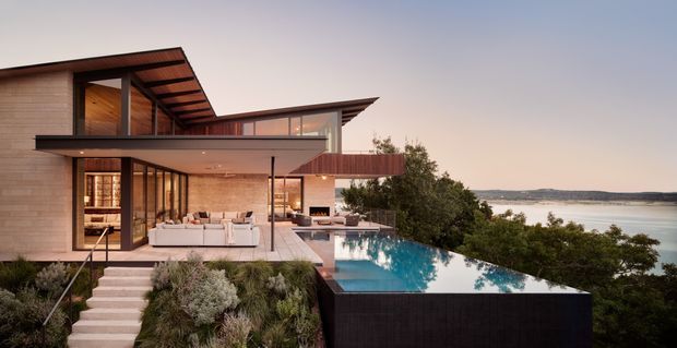 Modern multi-level waterfront residence with extensive timber cladding, broad overhanging roof planes, floor-to-ceiling glazing, and a cantilevered upper volume. A furnished terrace opens onto an infinity-edge swimming pool overlooking the water, with landscaped planting around the stepped approach.