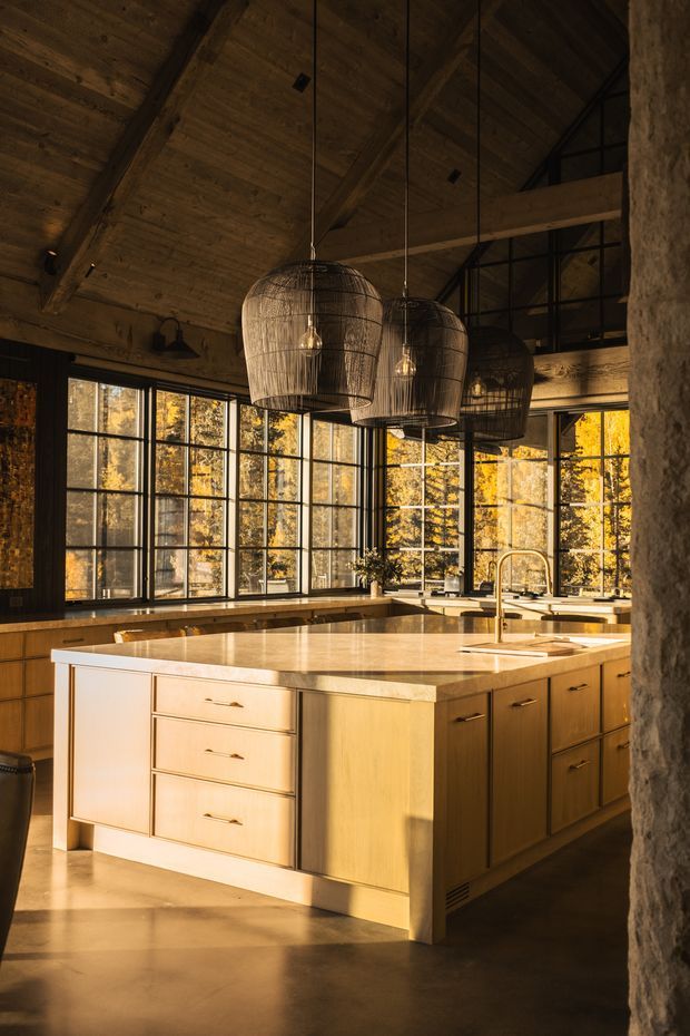 A spacious, warmly lit kitchen with a large central island, pale timber cabinetry, and a light stone benchtop. The room features extensive black-framed floor-to-ceiling glazing, a dramatic pitched ceiling clad in timber with exposed structural beams, rustic woven pendant lights, and dark timber wall finishes. Golden sunlight and forest views create a lodge-like atmosphere, while a textured stone-clad wall frames the right side.