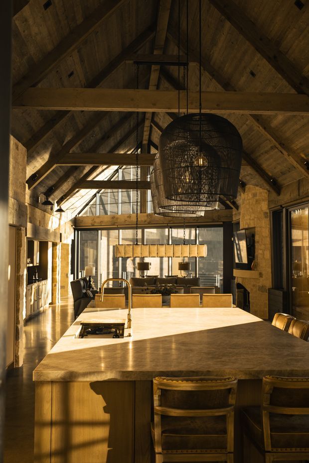 A warm, rustic kitchen inside a barn-like timber structure. The room features a large pale stone island with an integrated cooktop and brass-toned tap, timber cabinetry, exposed heavy timber beams and rafters, a pitched ceiling, dark woven pendant lighting, and full-height glazed doors or windows at the rear. The kitchen opens toward a dining and living area, with strong late-afternoon sunlight creating a golden, dramatic atmosphere.