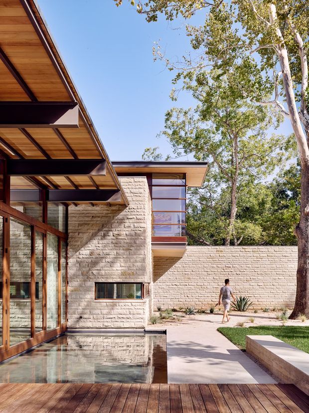 A modern multi-level residential courtyard viewed from a timber deck, featuring broad projecting roof eaves with exposed dark structural beams and timber soffits, extensive timber-framed glazing, pale textured masonry walls, and a shallow reflecting pool beside the deck. A paved courtyard with minimalist planting and mature trees extends around the building.