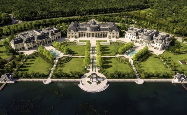 Aerial view of a grand symmetrical estate arranged around formal manicured gardens and a central axis leading to a broad waterfront. The large central mansion and flanking wings feature pale stone façades, dark steeply pitched roofs, dormer windows, classical proportions, ornate roof elements, and extensive formal glazing. Geometric lawns, clipped hedges, fountains or pools, axial pathways, and a semicircular waterfront terrace create a highly formal, palace-like composition.