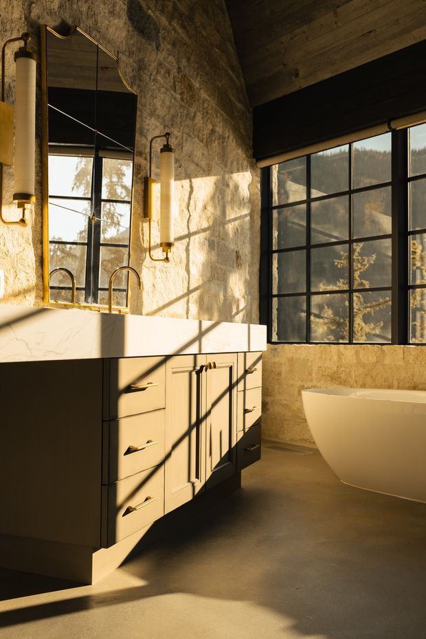 A warm, moody bathroom interior featuring a freestanding white bathtub beside a dark-framed industrial-style window. A large marble-look vanity with integrated backsplash, dark cabinetry, brass-toned pulls and twin wall-mounted cylindrical sconces sits beneath an irregular organic-textured wall and mirror. Timber ceiling boards and strong late-afternoon window shadows add warmth and drama.