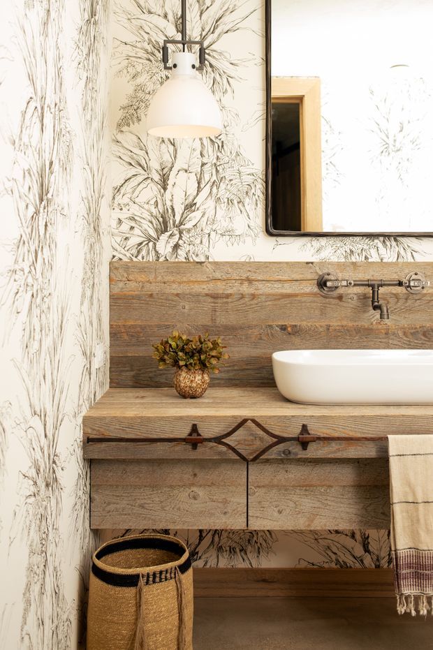 A warmly lit bathroom vanity vignette featuring a white vessel basin on a rustic reclaimed timber wall-mounted cabinet and benchtop. A vintage-style exposed wall-mounted tap, black metal pendant light, framed mirror, botanical illustrated wallpaper, woven basket, and striped hanging towel contribute to the rustic character.
