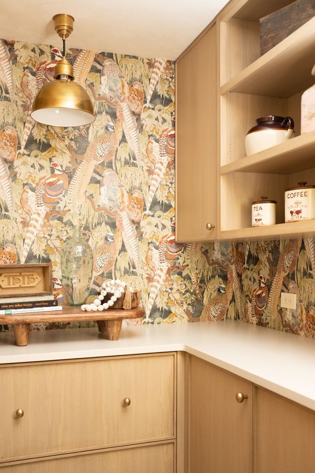 A warm, compact kitchen corner with light timber cabinetry, open shelving, and a white L-shaped benchtop. The walls are covered in a highly decorative botanical and bird-patterned wallpaper, complemented by a brass pendant light and vintage-inspired accessories, creating a colourful retro eclectic character.