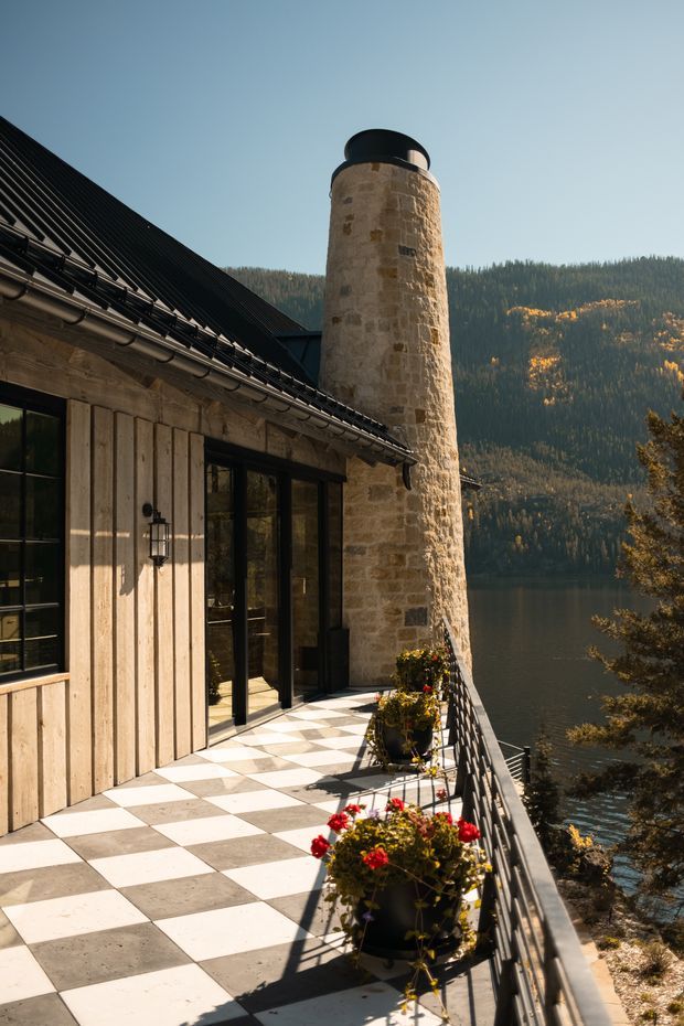 Exterior architectural view of a rustic lodge or residence beside a forested lake, showing a long balcony terrace with black-and-white checkerboard paving, black metal balustrade, vertical timber cladding, dark-framed glazing, and a prominent cylindrical stone chimney. Mountains and water are visible beyond the building.