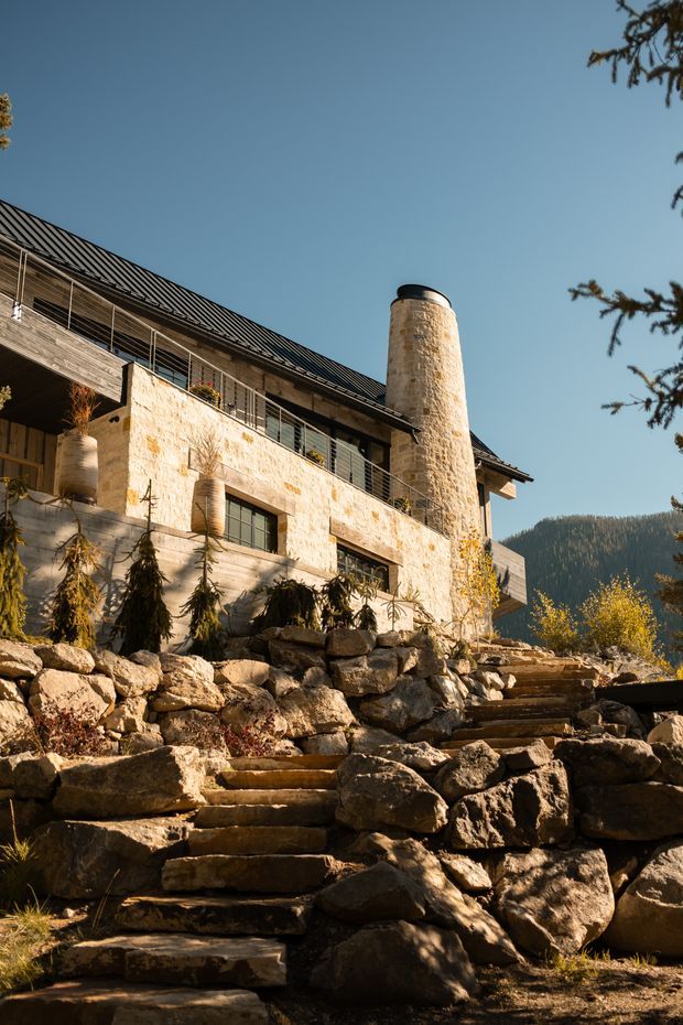 Exterior view of a multi-level stone and timber building set into a rocky, landscaped mountain site. The facade combines pale rough-cut stone walls, horizontal timber cladding, dark metal balcony railings, and a steep dark metal pitched roof. A prominent cylindrical stone chimney rises beside the building, while irregular stone retaining walls and steps lead up through the foreground. Forested mountain terrain is visible in the background.