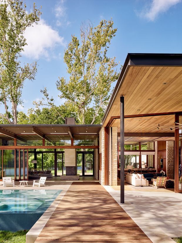 A high-end modern residential courtyard viewed from outdoors, centred on a rectangular swimming pool and broad timber decking. The house has extensive floor-to-ceiling glazing, open-plan indoor-outdoor living areas, exposed timber soffits and ceilings, dark slender metal framing, and warm brick walls. A covered outdoor lounge and living room open directly onto the pool terrace, surrounded by mature trees and bright sky.