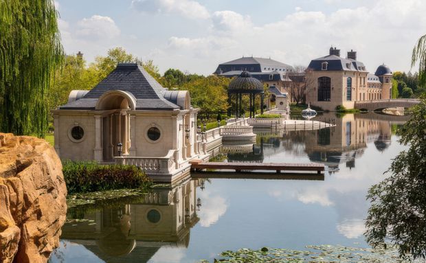 A grand European-inspired estate arranged around a calm ornamental lake, with ornate stone buildings, steep slate roofs, classical columns, balustrades, domed garden structures, and formal landscaped grounds. The architecture is reflected in the water, creating a stately, highly picturesque scene.