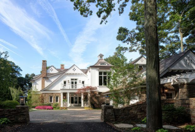 A large traditional residence viewed from a landscaped driveway, featuring multiple gabled roof forms, white painted siding, stone-clad lower walls, brick chimneys, decorative timber detailing, dormers, and a prominent central cupola. Mature trees, layered stone retaining walls, and garden planting frame the facade.