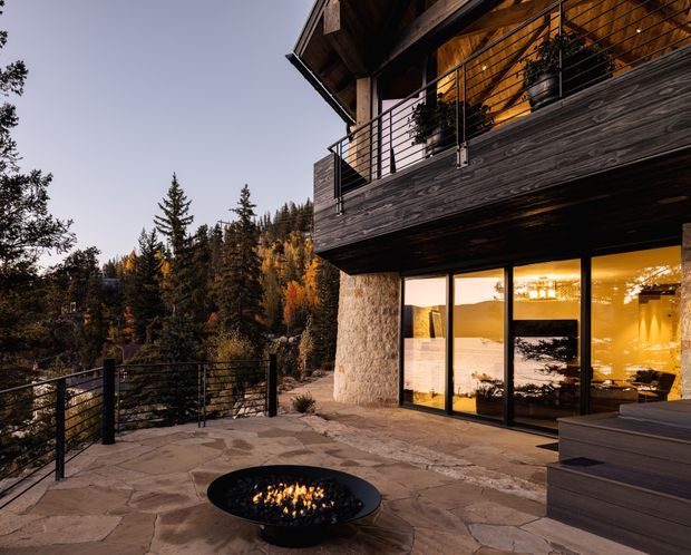 A contemporary mountain retreat viewed from a stone-paved outdoor terrace at dusk. The home features dark timber cladding, extensive floor-to-ceiling glazing, a projecting upper balcony with black metal cable railings, and a substantial natural-stone column. Warm interior lighting, a built-in outdoor fire bowl, and forested alpine surroundings create a secluded, inviting setting.