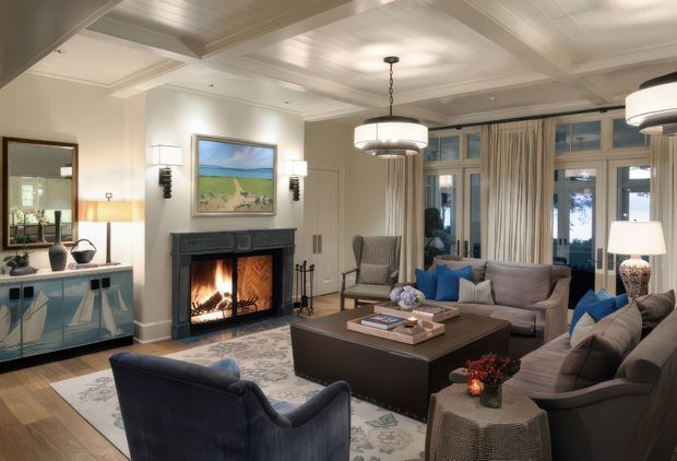 Elegant, traditionally styled living room with a coffered ceiling, detailed cornices, tall multi-pane windows with full curtains, and a dark fireplace surround with a lit fire. The room contains a large sectional sofa, upholstered armchairs, a substantial dark coffee table, side tables, lamps, a console, and a patterned area rug. The palette is warm and refined, combining soft neutral walls and upholstery with dark timber furnishings and blue accents.