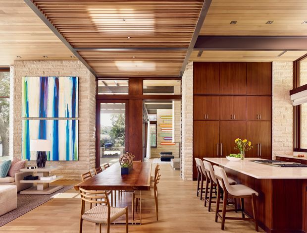 Open-plan contemporary interior combining a dining area and kitchen, with warm timber flooring, a slatted timber ceiling, extensive dark timber cabinetry, pale stone or brick-textured columns, and large glazed openings. A long dining table sits beside a kitchen island with upholstered bar stools. Colourful abstract artwork and restrained furnishings add visual contrast.