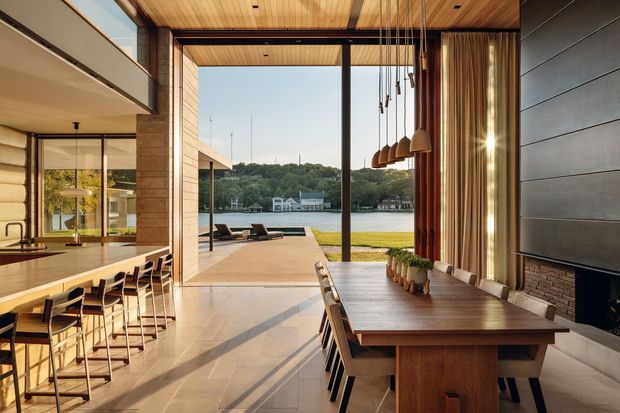A refined open-plan waterfront interior with a kitchen, dining area, and living space arranged around expansive floor-to-ceiling glazing. Warm timber ceilings, exposed structural framing, vertical timber screens, a long timber dining table, pendant lights, and a dark fireplace wall create a sophisticated connection to the outdoor terrace, lawn, and water beyond.