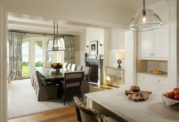 A bright, refined open-plan kitchen and dining area with a large pale stone island in the foreground, white built-in cabinetry, timber flooring, and a formal dining table beneath statement pendant lights. The dining area includes upholstered chairs, patterned full-height curtains, and a fireplace with dark surround; French doors open to a green outdoor view. The interior has classic panelled detailing with a calm neutral palette and traditional furnishings.