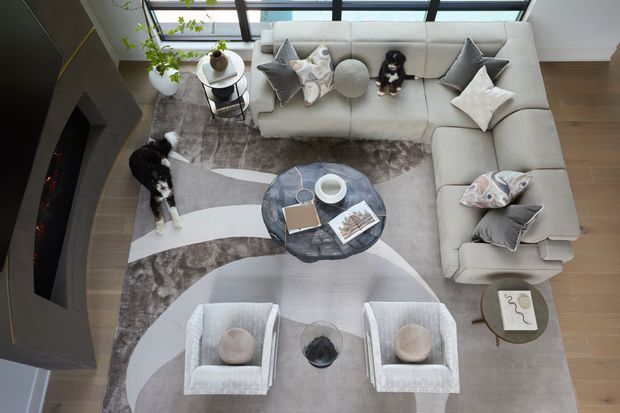Overhead view of a bright, refined living room arranged around a large pale sectional sofa. The room includes two upholstered lounge chairs, a round dark coffee table, layered neutral patterned rugs, decorative cushions, indoor plants, and a built-in dark fireplace along the left wall. Floor-to-ceiling glazing and a rooflight-like glazed opening bring in abundant natural light, while timber flooring and soft grey, white, and taupe furnishings create a calm contemporary atmosphere.