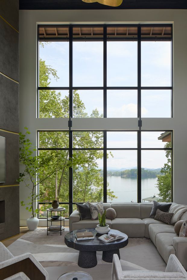A sophisticated living room with a double-height wall of black-framed grid windows overlooking a lake and wooded landscape. The room features a large light-grey sectional sofa, sculptural dark stone coffee table, upholstered armchairs, neutral layered rug, potted tree, and soft contemporary furnishings. The expansive glazing and high ceiling create a bright, airy connection to the outdoors.