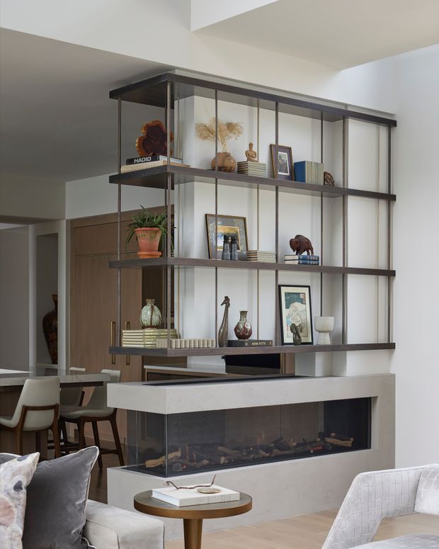 A contemporary open-plan living area centered on a built-in linear fireplace with a white stone or rendered surround and a dark glass firebox. A dark timber and metal open shelving unit rises above the fireplace, displaying books, framed artwork, ceramics, sculptures, and decorative objects. The room includes a soft neutral sofa and armchair, a small round timber coffee table, and a dining area visible behind the shelving. White walls, a high ceiling, timber flooring, and warm natural styling create a refined, inviting interior.