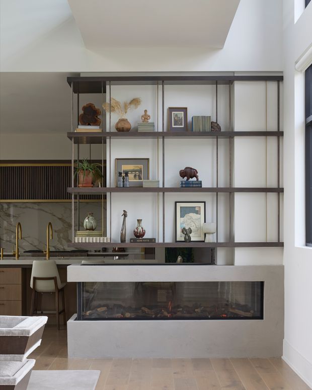 A refined open-plan living and kitchen area featuring a full-height dark metal and timber display shelving unit dividing the spaces. The shelving is styled with books, framed artwork, ceramics, sculptures, and plants, and incorporates a wide built-in fireplace with a pale stone or plaster surround and glazed firebox. A marble-look kitchen splashback, timber cabinetry, and brushed brass tapware are visible behind the shelving. The room has light flooring, white walls, high ceilings, and abundant natural light.