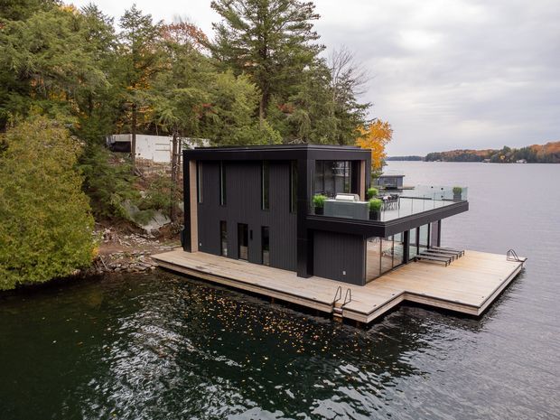 A modern dark-clad floating or waterside pavilion built on a large timber deck over a calm lake. The compact two-storey structure has a flat roof, vertical dark cladding, extensive glazing on the waterside, an upper terrace with outdoor furniture, and a small dock with a ladder. Dense mature woodland surrounds the rocky shoreline, with autumn foliage visible across the lake.