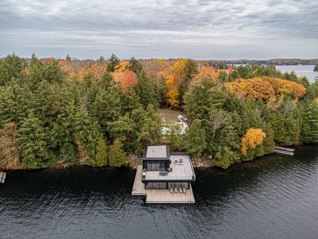 Aerial view of a compact modern waterfront house or pavilion projecting over a dark lake, surrounded by dense mixed forest in vivid autumn colours. The building has a flat roof, extensive glazing, a rooftop or upper terrace, and a timber deck with a small dock at the water's edge.