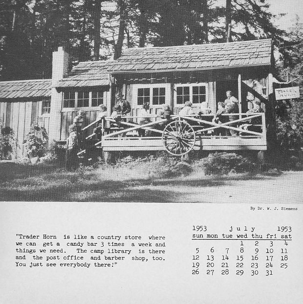 A black-and-white archival photograph of a rustic timber country store or trading post in a forested setting. The small single-storey building has a shingle-clad pitched roof, chimney, timber walls, multi-pane windows, and a covered veranda with crossed timber railing. A large wagon wheel is mounted on the front railing, and several people sit or stand on the porch. A printed caption identifies the building as “Trader Horn,” a country store also containing a café, library, post office, and barber shop, with a July 1953 calendar below.