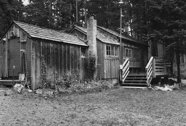 Black-and-white archival photograph of a rustic single-storey timber cabin or lodge in a wooded setting. The building has weatherboard walls, a shingled pitched roof with multiple gabled sections, timber-framed windows, a prominent masonry chimney, and a small raised entrance porch with steps and simple railings.