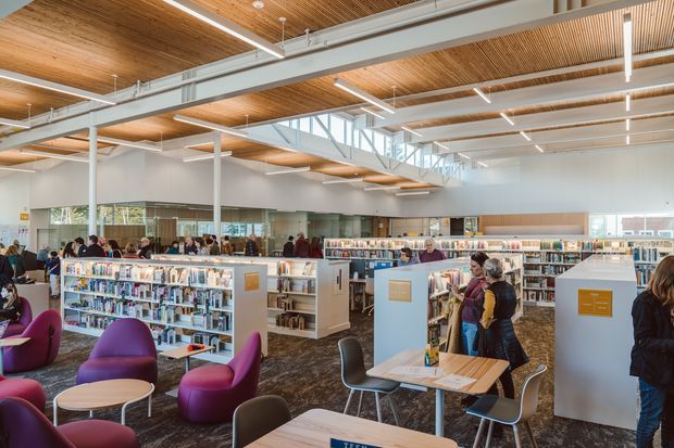 A bright, spacious contemporary library interior with open-plan book stacks, reading and study tables, lounge seating, and patrons browsing. The room features a high ceiling with exposed timber slatted panels, white-painted structural beams and columns, clerestory glazing, linear pendant lighting, and dark patterned carpet.