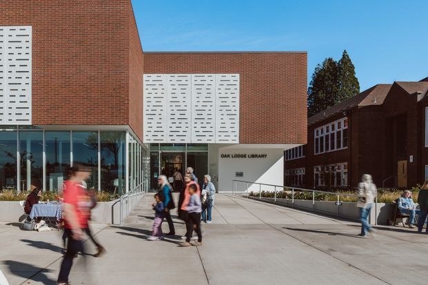 Exterior view of the Oak Lodge Library, a contemporary institutional building with a red brick facade, large glazed entrance, and distinctive white perforated screen panels. The library opens onto a broad concrete pedestrian forecourt, with an adjoining older brick building visible to the right.