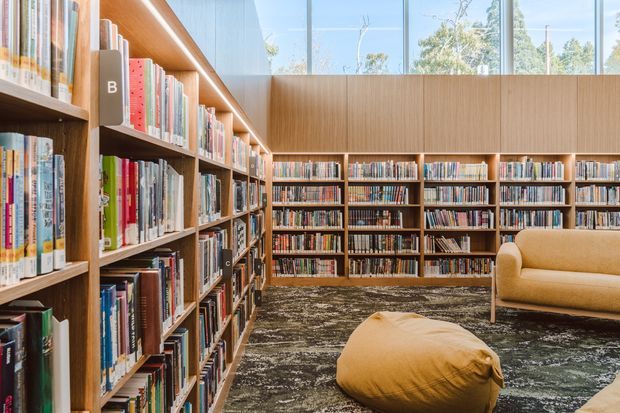 A bright, contemporary library interior with floor-to-ceiling timber bookshelves filled with books, large clerestory-style glazing, and a warm mustard upholstered sofa and floor cushion on a patterned dark carpet. The space feels inviting and thoughtfully designed, with abundant natural light and extensive built-in storage.