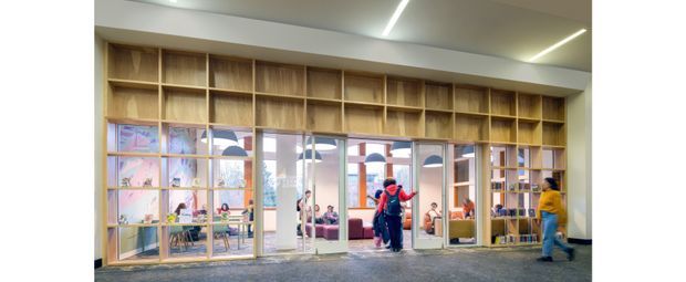 Bright contemporary commercial interior with a full-height timber display and shelving wall framing glazed openings into a colourful, active space beyond. Built-in open shelving contains books and objects, while linear recessed ceiling lights illuminate a polished concrete-like floor. Several people are visible, including a person walking through the foreground, and the glazing reveals additional seating and work or activity areas.