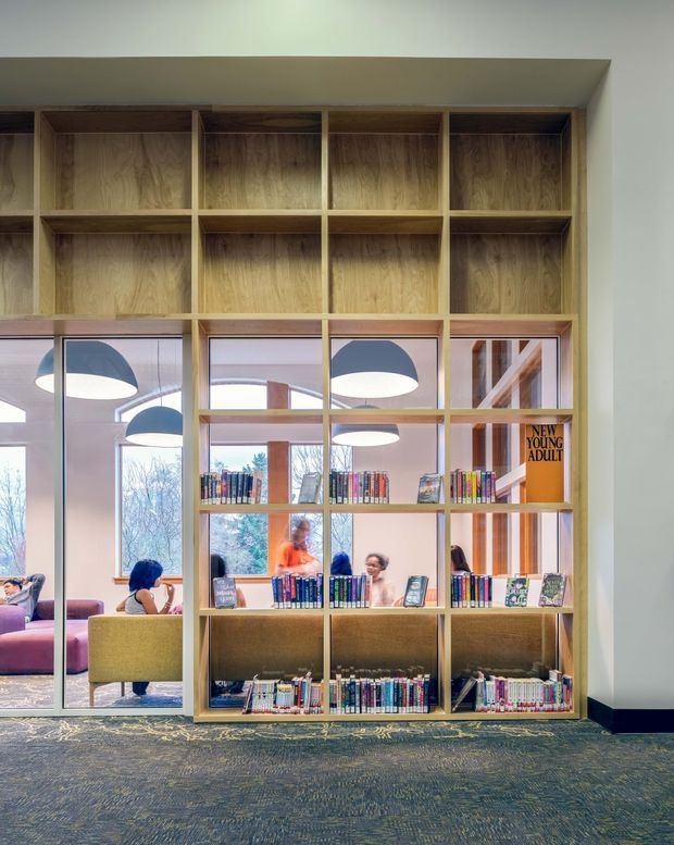A contemporary library interior featuring a large floor-to-ceiling timber bookshelf and room divider. The shelving combines enclosed wood-backed compartments above with open display shelves below, filled with books and visible library users. Through the lower open sections, a bright reading area with colorful upholstered seating, pendant lights, large windows, and glazed doors is visible. The space has patterned dark carpet, warm timber joinery, and a clean institutional design.