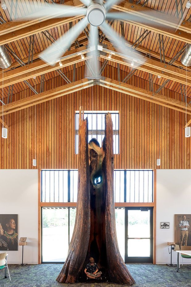 A tall, airy interior with a dramatic exposed timber roof structure, vertical timber wall panelling, clerestory-style windows, and large glazed doors. A monumental hollow timber sculpture dominates the centre of the space, which appears to function as a gallery or institutional display area. A large ceiling fan, suspended linear lighting, artwork, and carpeted flooring are visible.