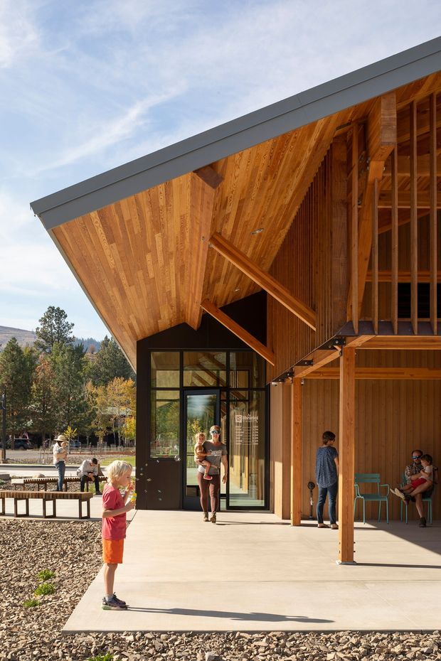 A modern timber-clad pavilion or community building viewed from a covered outdoor terrace. The structure features a dramatically sloping roof with deep eaves, exposed timber soffits and rafters, vertical timber screens, large dark-framed glazed doors, and an open unfinished-looking upper structural bay. People occupy the concrete terrace, with landscaped trees and hills visible in the background.