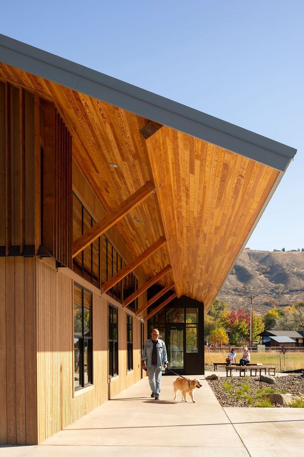 Exterior architectural photograph of a contemporary timber-clad building with a dramatic deep sloping roof canopy and exposed timber soffit and rafters. A covered concrete walkway runs along glazed and timber-clad walls, with a mountainous, semi-rural landscape visible beyond.