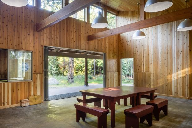 A spacious, high-ceilinged timber-lined dining and gathering room with exposed structural beams, polished concrete flooring, and a long solid timber table surrounded by matching benches and stools. Large operable glazed openings connect the room to a wooded garden, while multiple suspended dome lights provide illumination. The interior has a warm, natural, rustic character with strong indoor-outdoor connection.