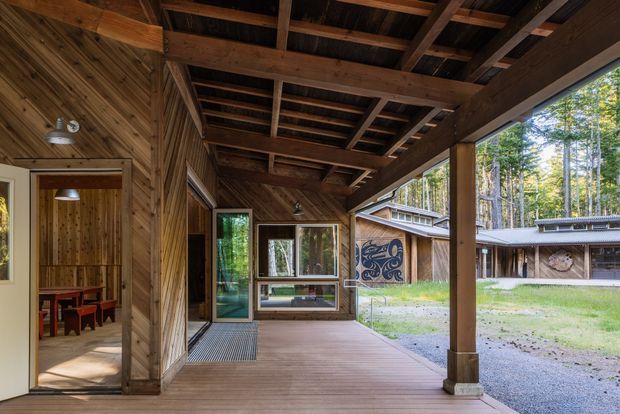 Covered timber-framed outdoor veranda or porch looking through a glazed opening toward a landscaped courtyard and additional low buildings. The structure features extensive dark-stained exposed timber posts, beams, rafters and diagonal wall cladding, with timber decking, large windows and a partially visible interior dining area. The setting has a rustic cabin-like character with strong connection to the surrounding wooded site.