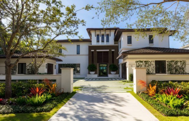 Front exterior view of a large, symmetrical white rendered residence with a central raised entry, dark timber accents, tiled low-pitched roofs, and landscaped garden beds. A broad paved pathway leads through manicured planting to the recessed front door, with mature trees framing the property.