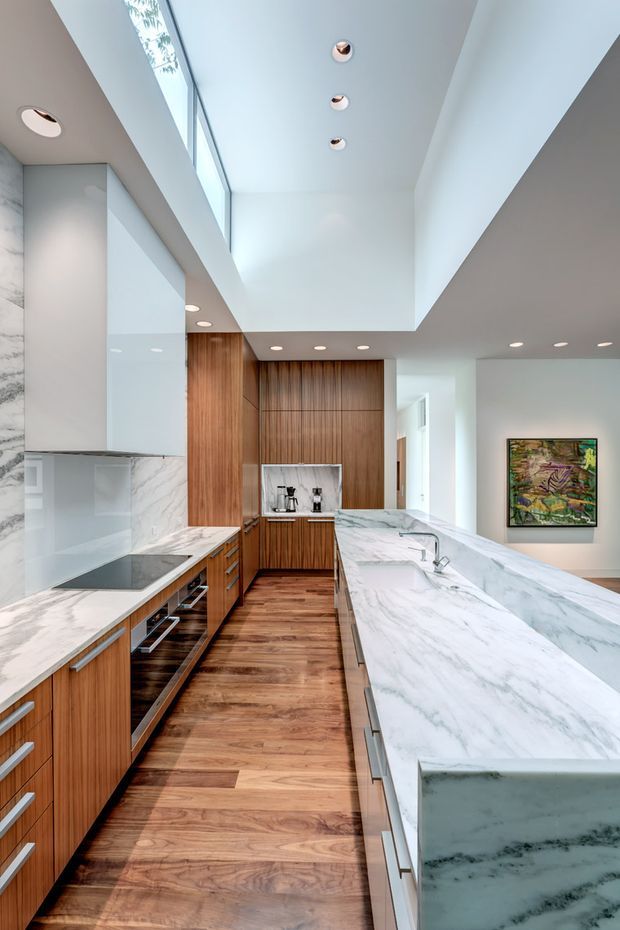 Bright contemporary kitchen with extensive warm timber cabinetry, white veined marble surfaces, a large waterfall island, integrated appliances, and a long skylight or clerestory opening above. Recessed downlights and a framed artwork visible beyond the kitchen contribute to the refined modern fit-out.
