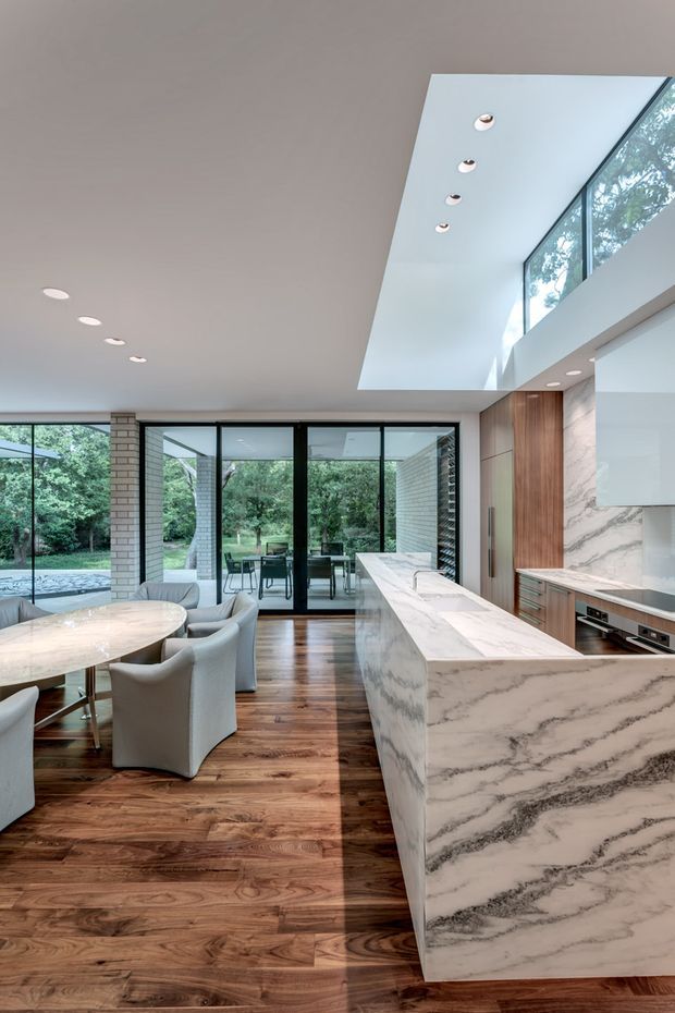 A bright, spacious contemporary kitchen and dining area with a large marble or engineered-stone island, integrated cabinetry, timber wall detailing, dark timber flooring, and extensive black-framed glazing. The room opens visually to a leafy outdoor garden and includes a dining table with upholstered chairs, recessed ceiling lighting, and a dramatic double-height skylit void.