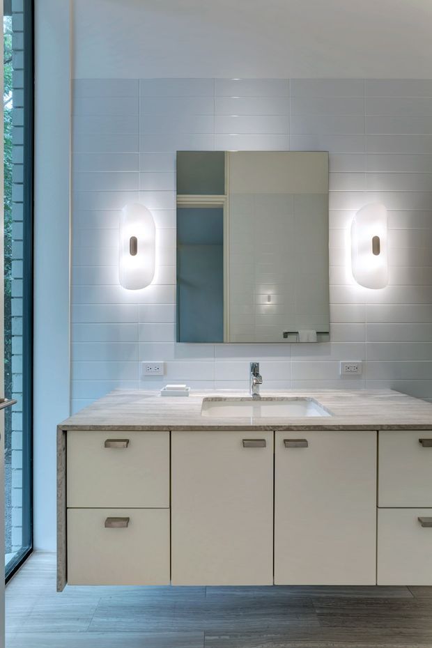 A contemporary bathroom vanity with a wide floating light-coloured cabinet, integrated rectangular basin, pale stone-look benchtop, large mirrored wall cabinet, and twin rounded wall sconces. The walls are clad in glossy white horizontal rectangular tiles, with dark timber-look flooring and a tall glazed opening at the left.
