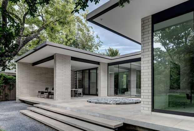 A contemporary single-storey courtyard-style residence with crisp white brick walls, flat roof planes, expansive dark-framed glazing, and a broad paved terrace. Stepped concrete access leads to the outdoor living area, which includes a partially covered seating zone and a round stone-lined spa or plunge pool. Mature trees surround the building and are reflected in the large windows.