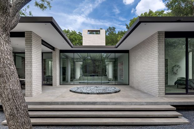 A contemporary single-storey courtyard residence viewed from the landscaped exterior. Pale brick wings with crisp flat rooflines frame a glazed central living space and a raised circular water feature. Broad stepped paving leads up to the terrace, while mature trees surround the house.