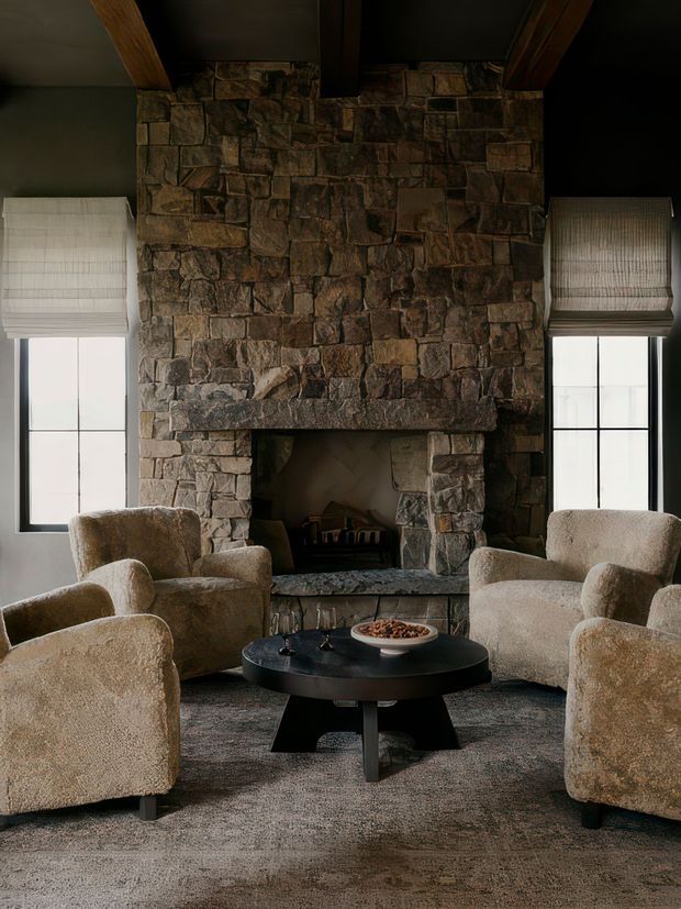 A moody, symmetrical living room centred on a large rustic stone fireplace with exposed timber ceiling beams. Two tall windows with Roman blinds flank the fireplace, while several rounded upholstered lounge chairs surround a low black coffee table on a textured grey carpet. The palette is earthy and warm, with substantial natural stone and soft neutral furnishings.