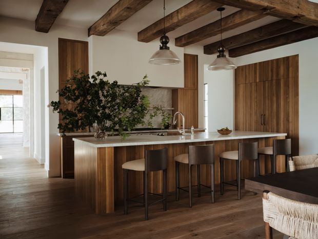 Warm, refined kitchen and dining area with a long wood-panelled island and integrated seating for five, dark timber ceiling beams, wood cabinetry, stone benchtops, pendant lights, and wide-plank timber flooring. A dining table and woven dining chairs are visible in the foreground, with an adjoining room beyond the opening.