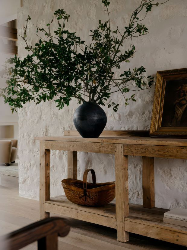 A warmly lit interior vignette featuring a rustic timber console table against a heavily textured white plaster wall. The console displays a large black ceramic vessel with leafy branches, a framed artwork, and a woven basket on the lower shelf. Light timber flooring and a partially visible upholstered chair contribute to the natural, refined character.