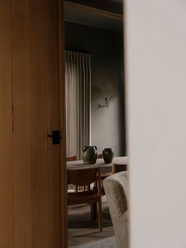 A dimly lit, refined interior viewed through a partially open timber door. The room contains a compact dining or work table with wooden chairs, ceramic vessels, a textured upholstered chair in the foreground, dark painted walls, sheer curtains, and a sculptural wall light. The palette is warm and earthy with strong timber detailing and soft, atmospheric lighting.