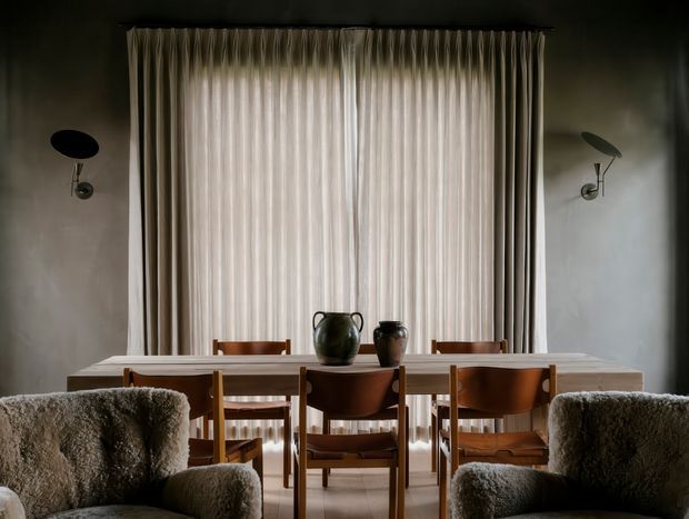 A moody, refined dining area viewed from an adjoining living space. A long light-toned dining table is surrounded by warm timber and brown leather chairs, with ceramic vessels as a simple centrepiece. Floor-to-ceiling sheer curtains cover the central window, while sculptural dark wall sconces flank it. Plush textured lounge chairs frame the foreground.