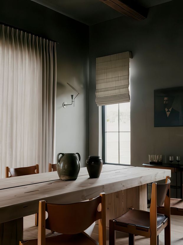 A moody, refined dining room featuring a long pale timber dining table, sculptural dark ceramic vessels, and leather-and-timber dining chairs. Dark charcoal walls, full-height sheer curtains, a woven Roman blind, minimalist wall lighting, and a tall divided window create a warm, architectural atmosphere.