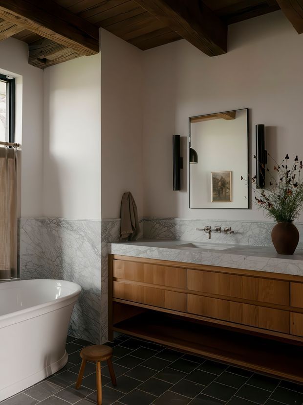 A refined bathroom featuring a freestanding white bathtub, a long timber vanity with integrated basins, a pale veined marble wall and benchtop finish, dark tiled flooring, and a large wall mirror. Exposed dark timber ceiling beams and boards add warmth and rustic character, while black wall-mounted fixtures, a stool, and a sculptural vase provide contrast.